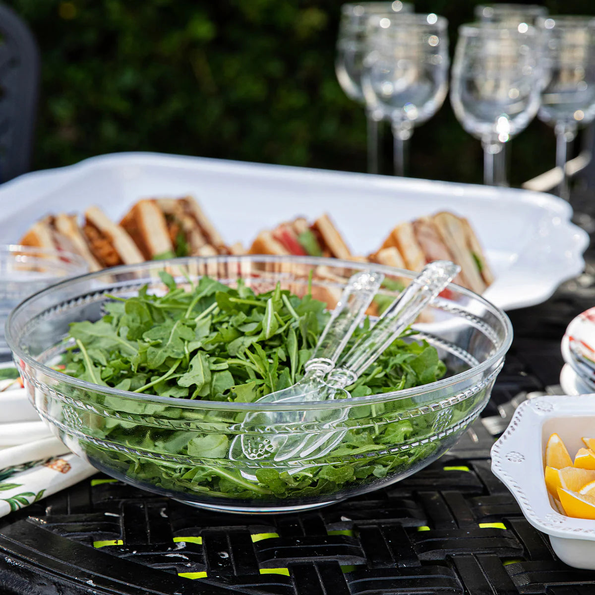 Isabella Acrylic Salad Server Outdoor setting with a bowl of greens, sandwiches, and wine glasses on a table.