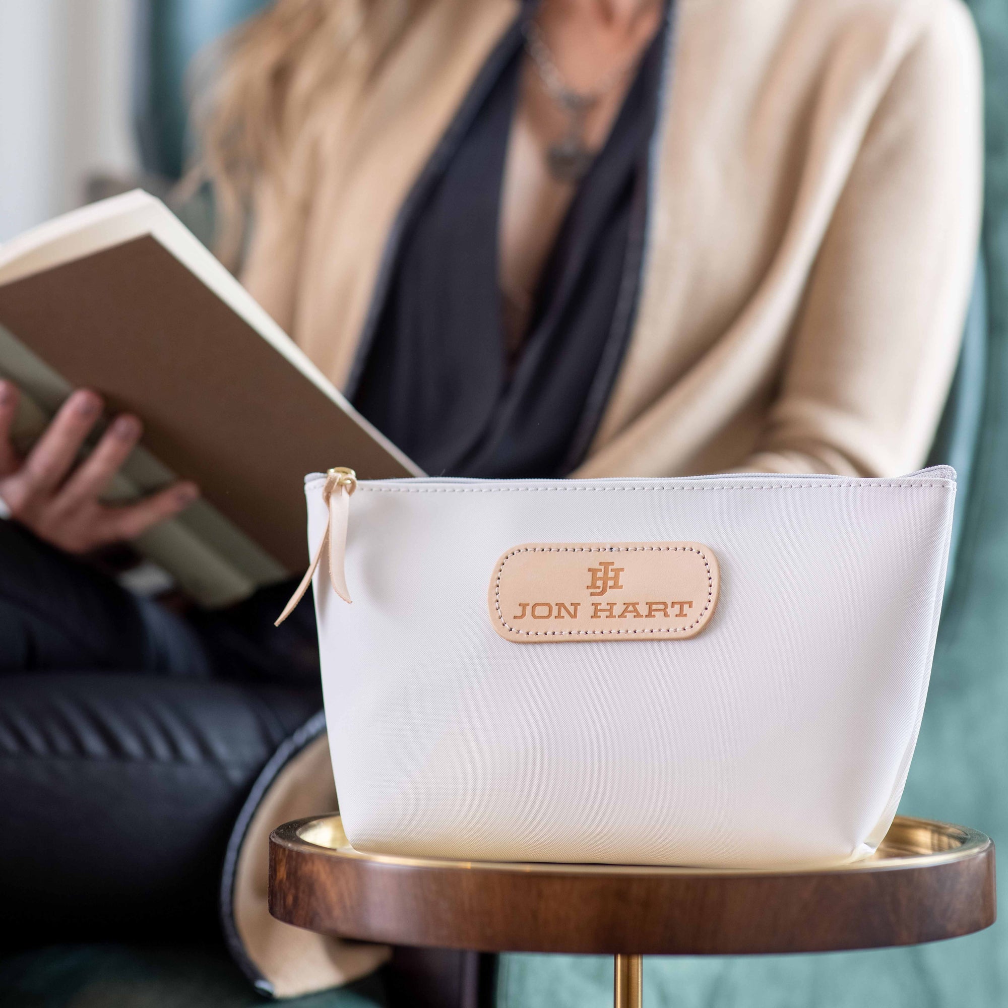 A white canvas cosmetic bag with natural leather trim and the brand 'JON HART' visible on it, displayed on a wooden table with a person sitting behind it reading a book.