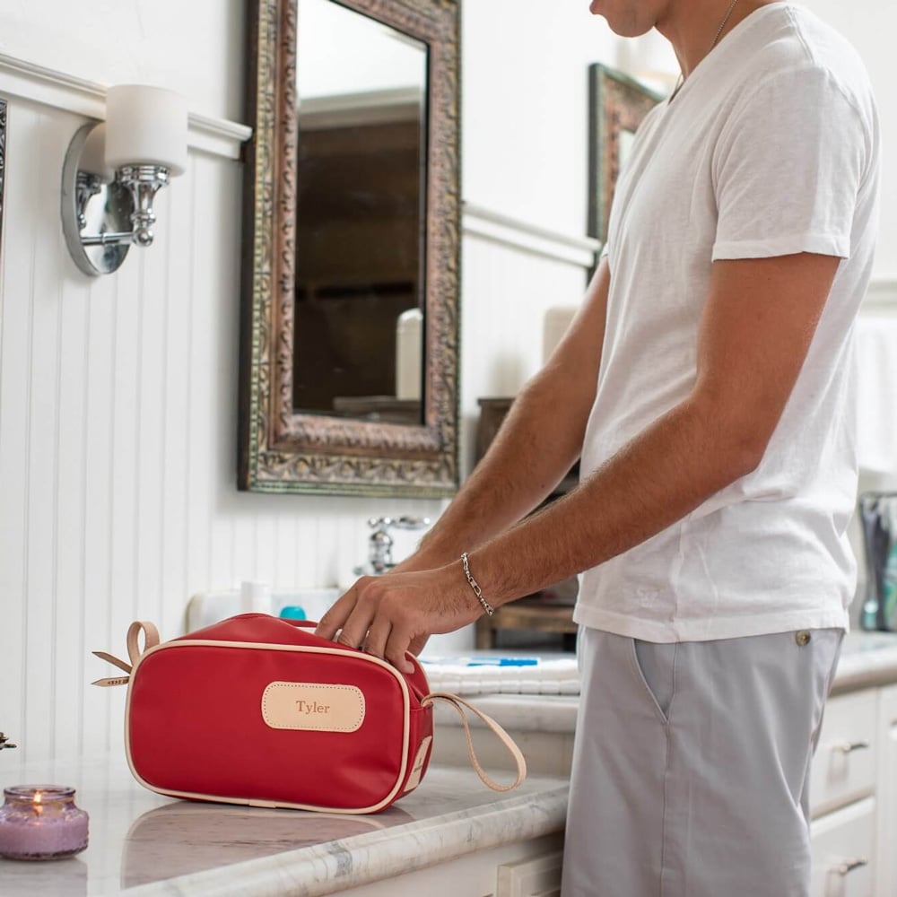 A man standing in a bathroom with a red and natural leather shaving kit bag placed on the countertop.